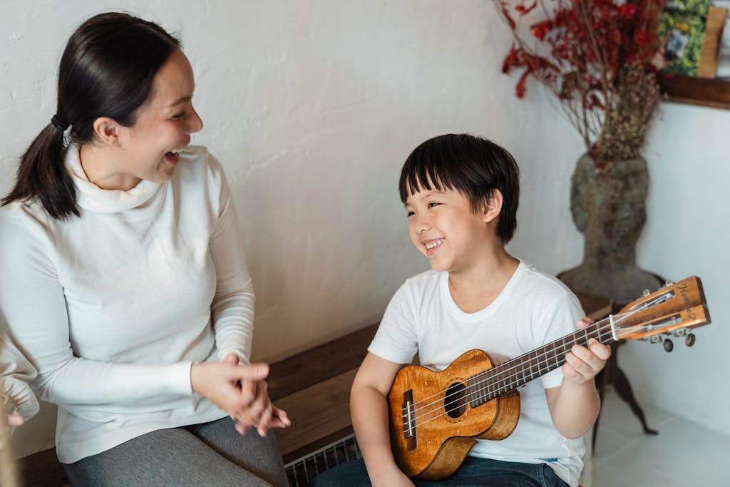 Positive ethnic child sitting with traditional Hawaiian musical instrument and looking away near content mother sitting with open mouth near vase with decorative branch
