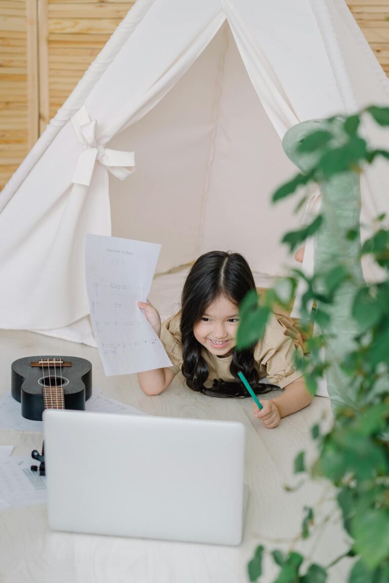 A smiling child engages in music and online learning indoors with a ukulele and laptop.